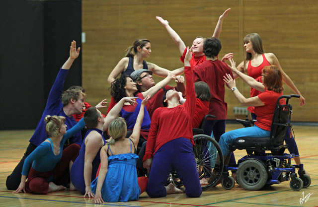 Picture of a large group of CRIPSiE dancers in blue and red all crowded together and reaching towards one dancer in the middle. Some dancers are standing, some kneeling, some sitting in wheelchairs,